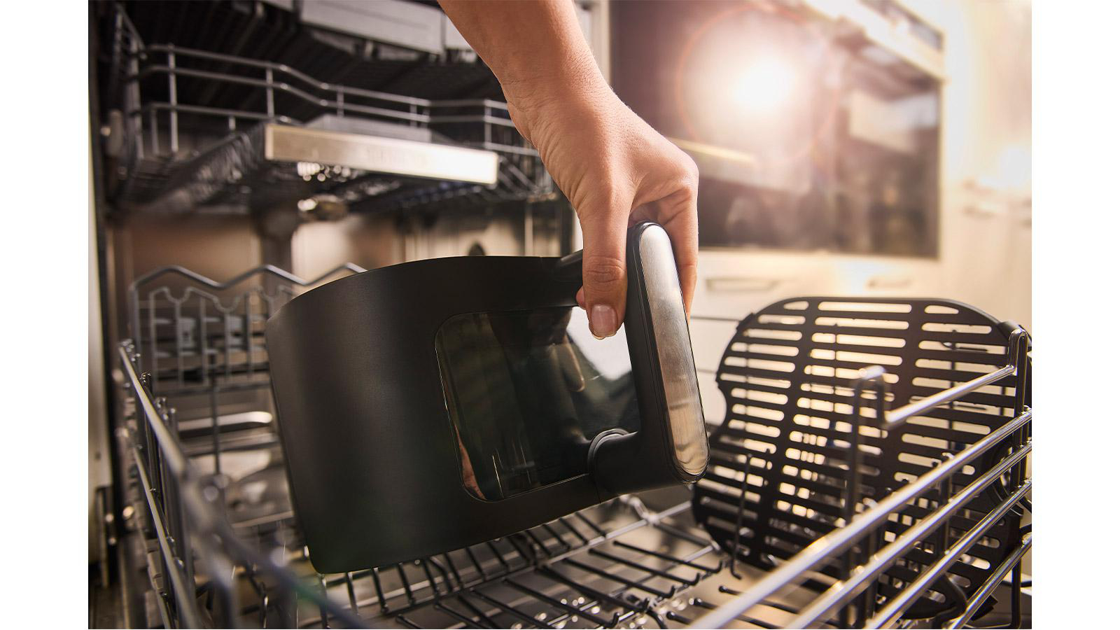 Eine Hand legt einen schwarzen Airfryer-Korb in einen Geschirrspüler. Der Korb wird über den Metallgestellen gehalten. Helles Licht scheint.