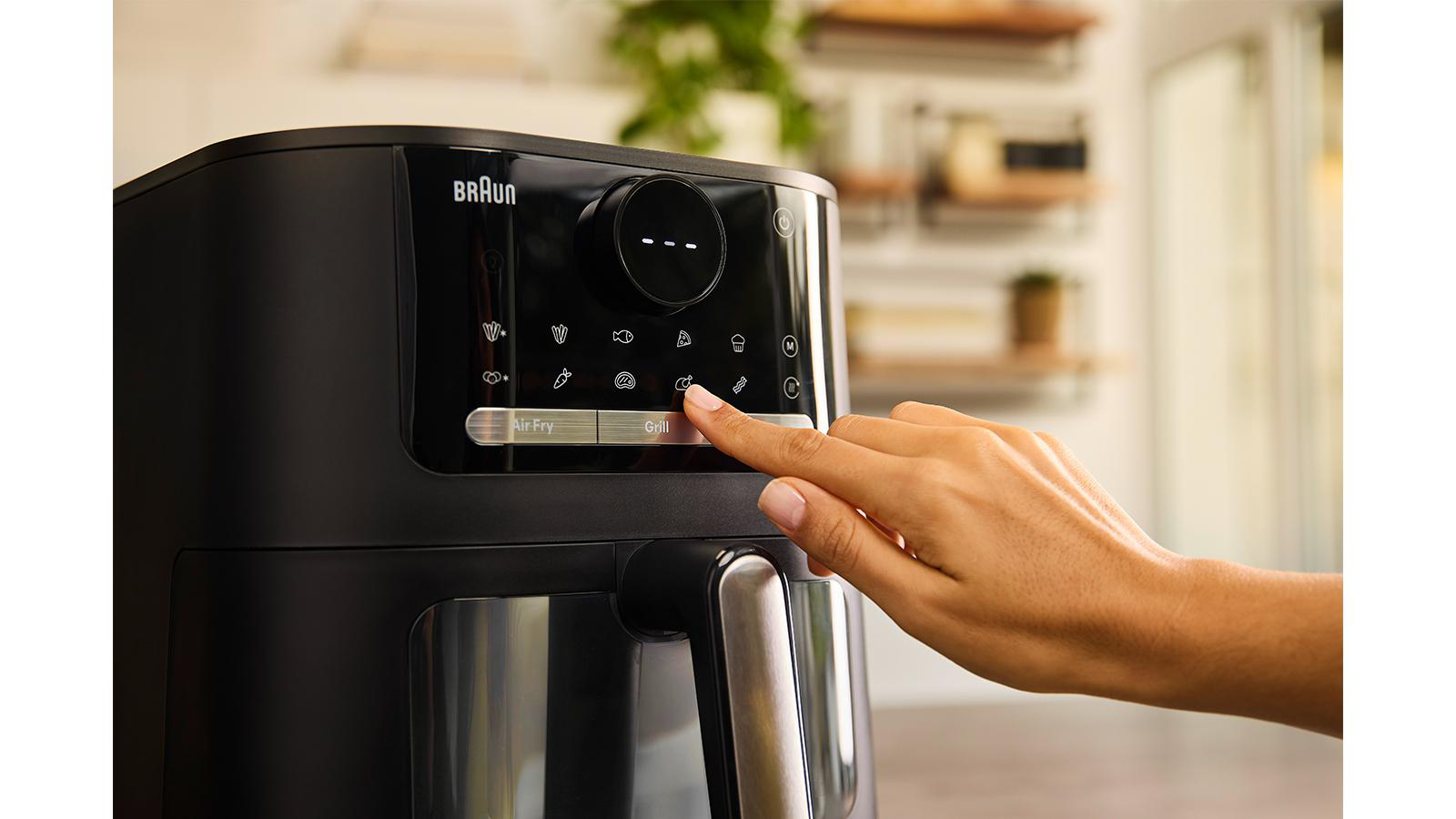 Eine Hand drückt die Taste 'Grill' auf einem schwarzen Braun-Heißluftfritteuse. Der Hintergrund zeigt eine Küche mit Regalen und Pflanzen.