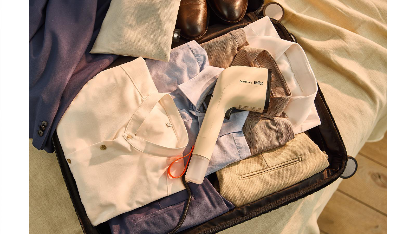 Ein Koffer ist geöffnet, gepackt mit Kleidung und einem Braun-Dampfgarer. Es gibt Hemden, Hosen und Schuhe. Der Hintergrund ist eine beige Oberfläche.