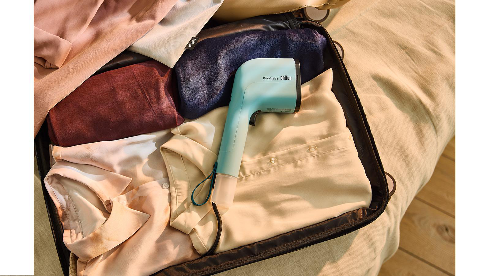Ein offener Koffer voller Kleidung und ein blauer Kleiderdampfer. Die Kleidung ist in verschiedenen Farben gehalten. Der Koffer steht auf einem Bett.