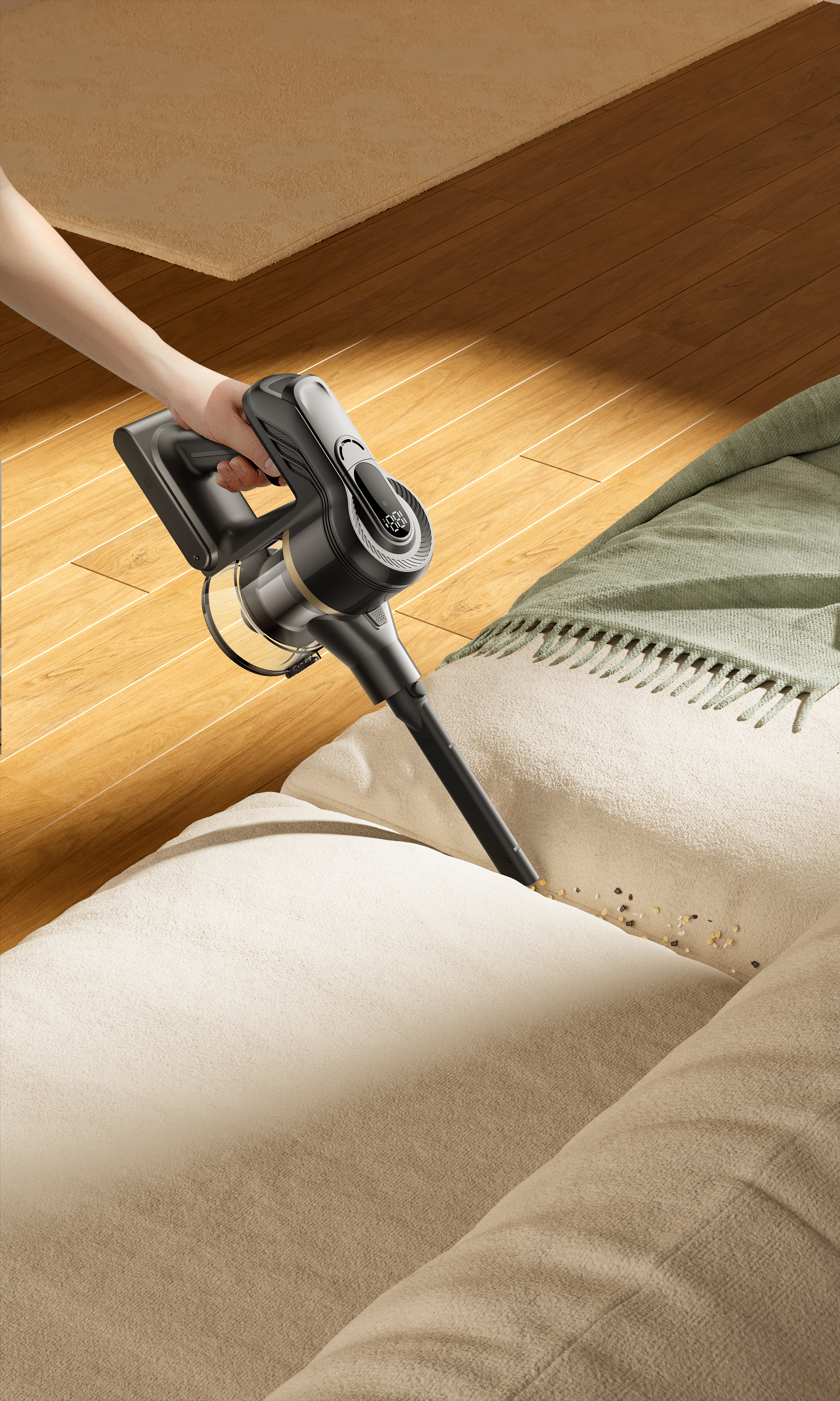 Ręka trzyma czarno-szary odkurzacz nad beżową sofą z zanieczyszczeniami. W pobliżu zielony koc na drewnianych podłogach.