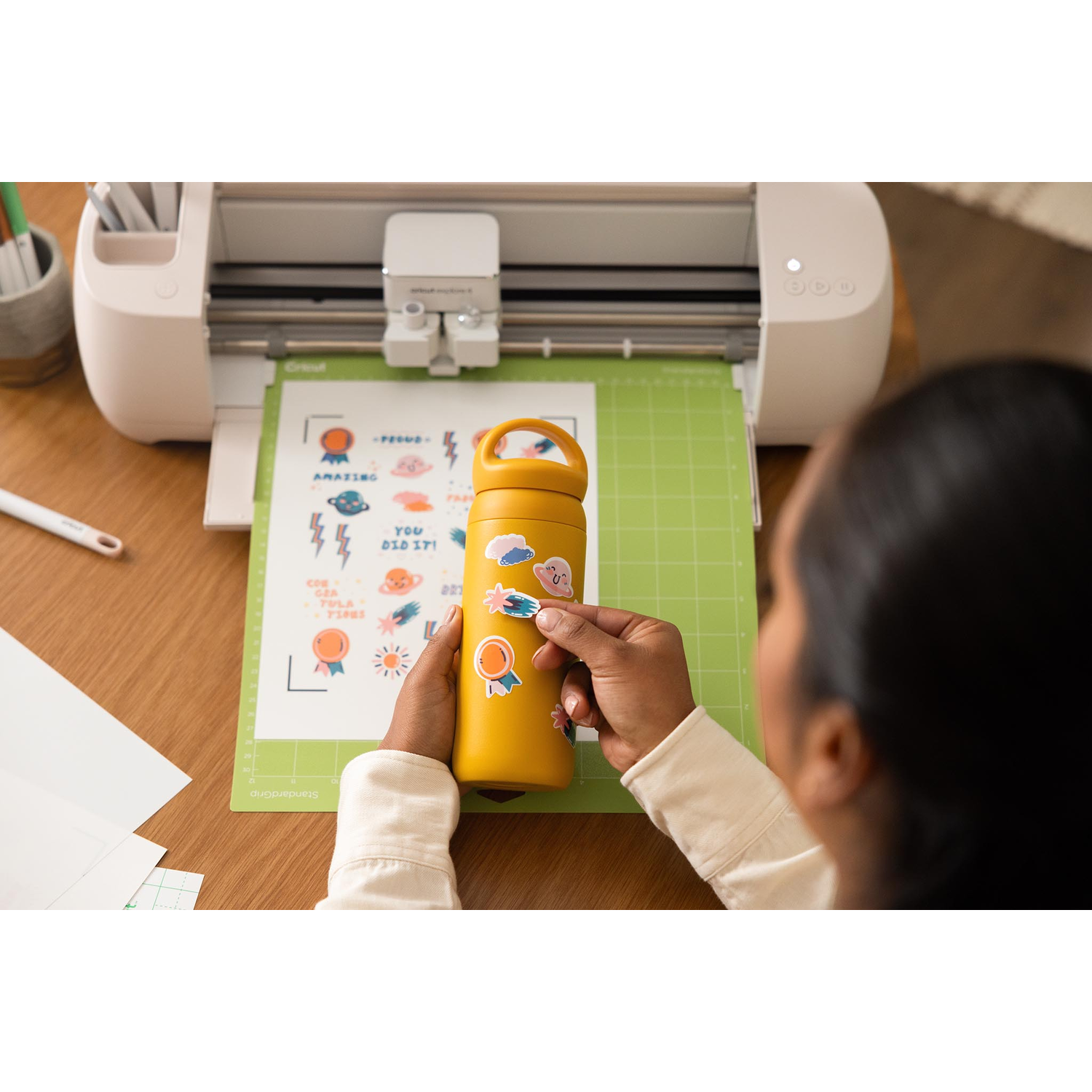 A person applies stickers to a yellow water bottle. A Cricut machine sits in the background with a green cutting mat.
