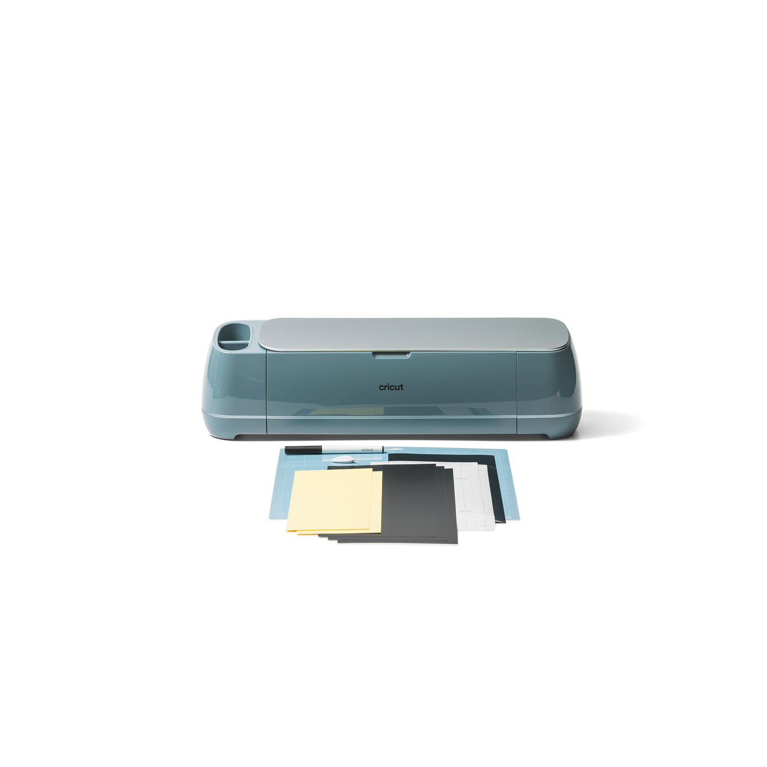 A teal Cricut machine sits on a white surface, with various colored paper sheets in front. The background is white.