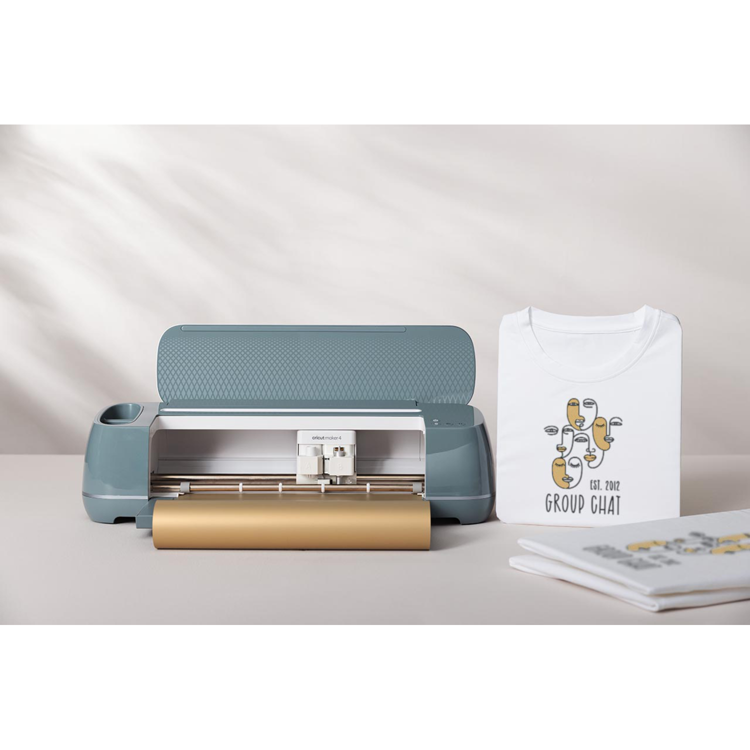 A Cricut Maker 4 machine with a gold roll, a white t-shirt with a logo, and another folded t-shirt are on a white surface.