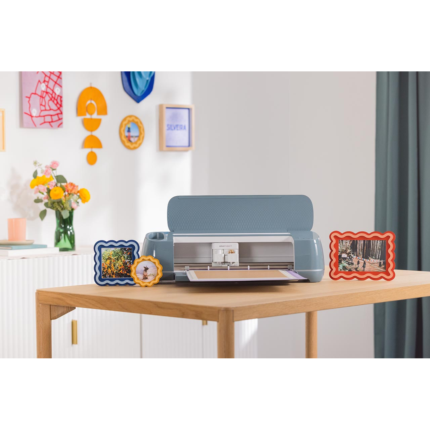 A blue crafting machine sits on a wooden table with picture frames. Colorful wall decorations and flowers are in the background.