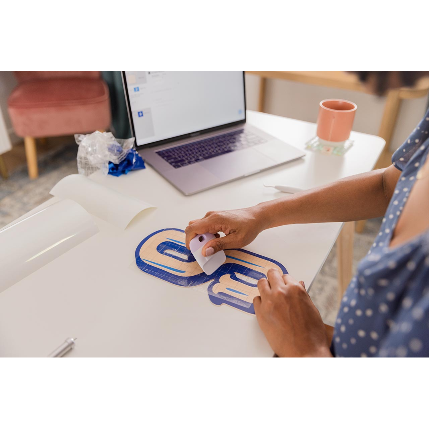 Person using a purple tool to apply vinyl letters on a white table. A laptop, cup, and chair are in the background.