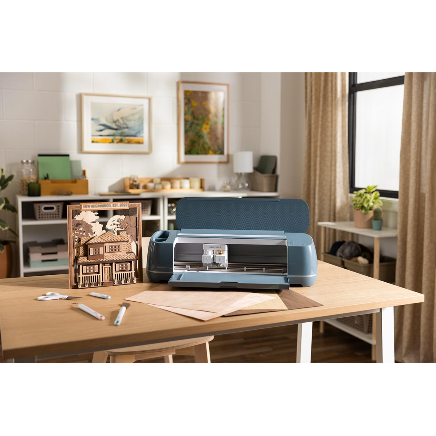 A blue Cricut machine sits open on a wood table. A wooden house art piece and art supplies are on the table.