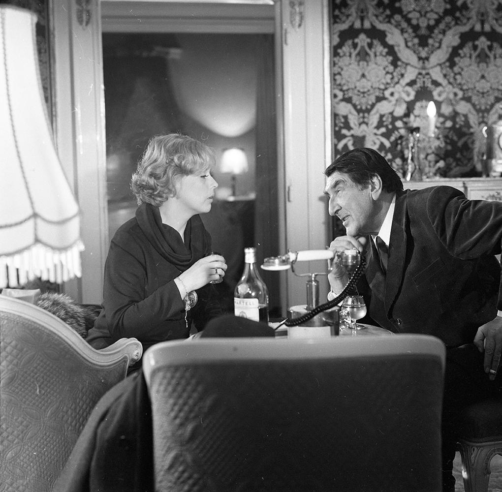 Eine Frau und ein Mann sitzen an einem Tisch. Der Mann hält einen Telefonhörer. Auf dem Tisch steht eine Flasche Alkohol.