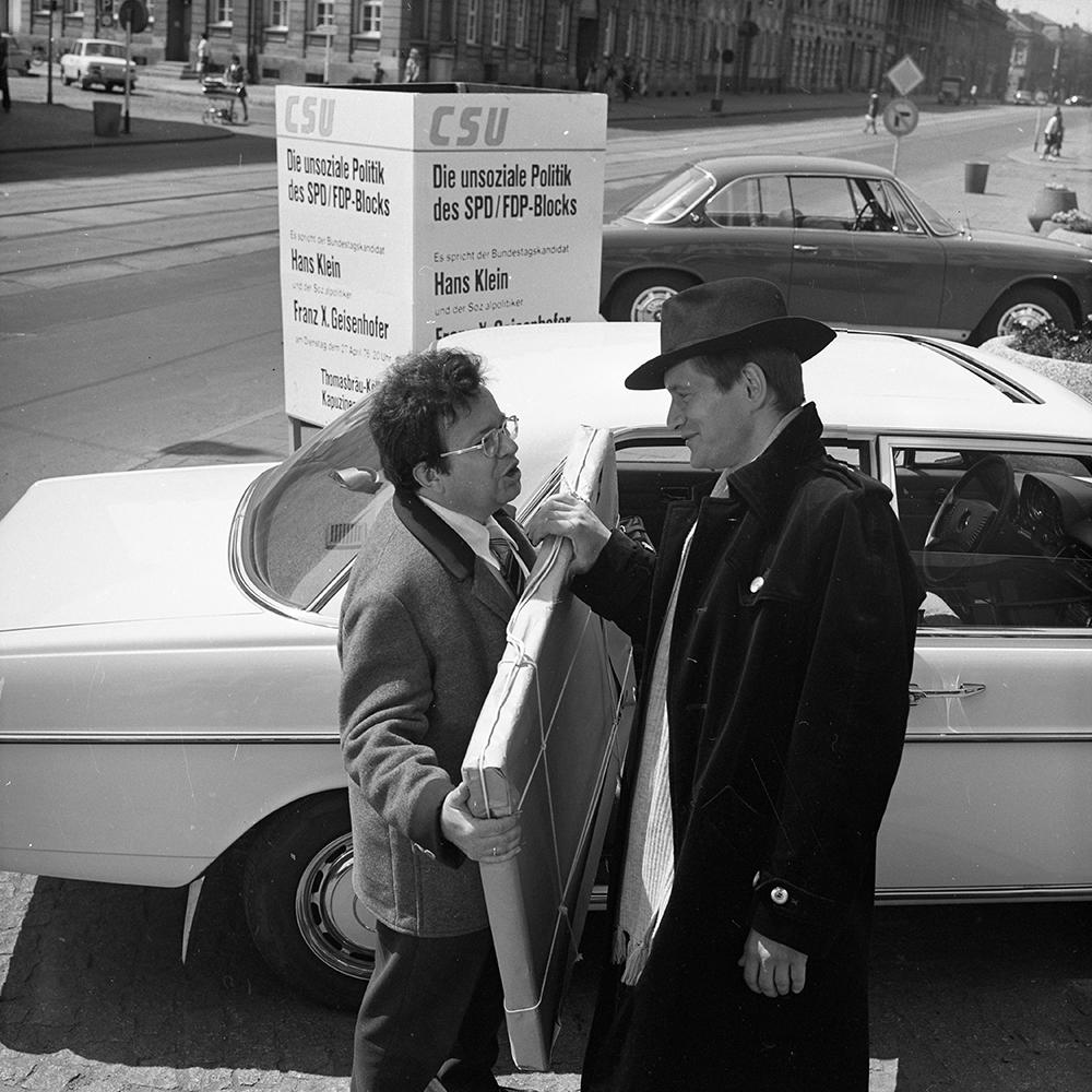 Zwei Männer an einem Auto, einer hält ein Schild mit der Aufschrift CSU. Autos und Gebäude im Hintergrund. Schwarz-Weiß-Foto.