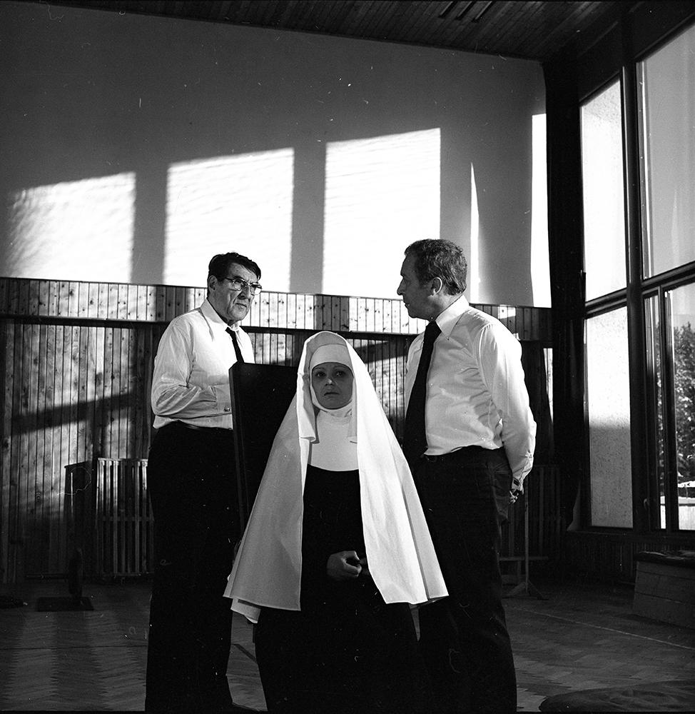 Eine Nonne in weißem Habit sitzt zwischen zwei Männern in weißen Hemden und schwarzen Krawatten. Sonnenlicht fällt durch große Fenster.