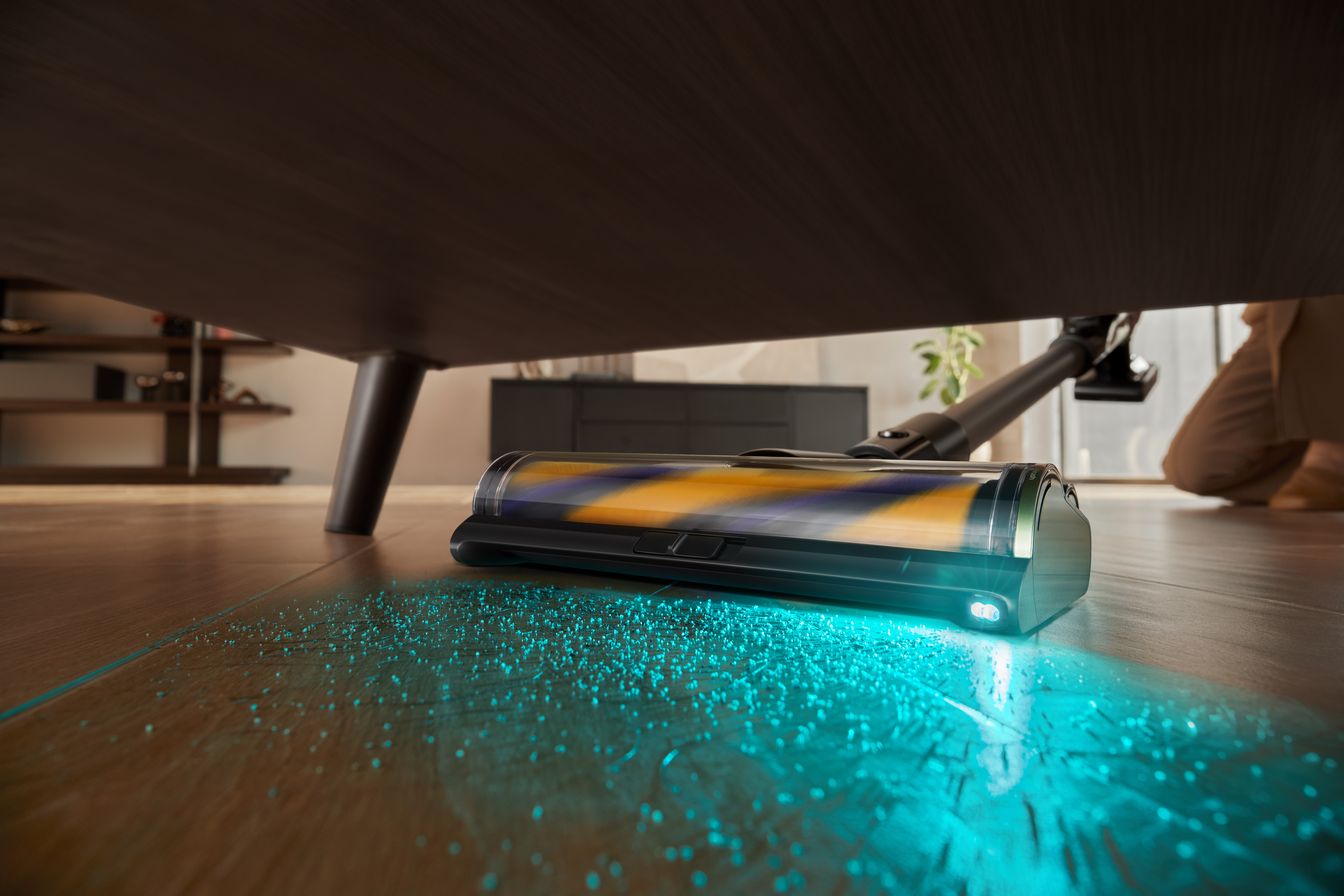 Odkurzacz czyści drewnianą podłogę pod stołem. Odkurzacz ma światło i usuwa zanieczyszczenia. Osoba klęczy.