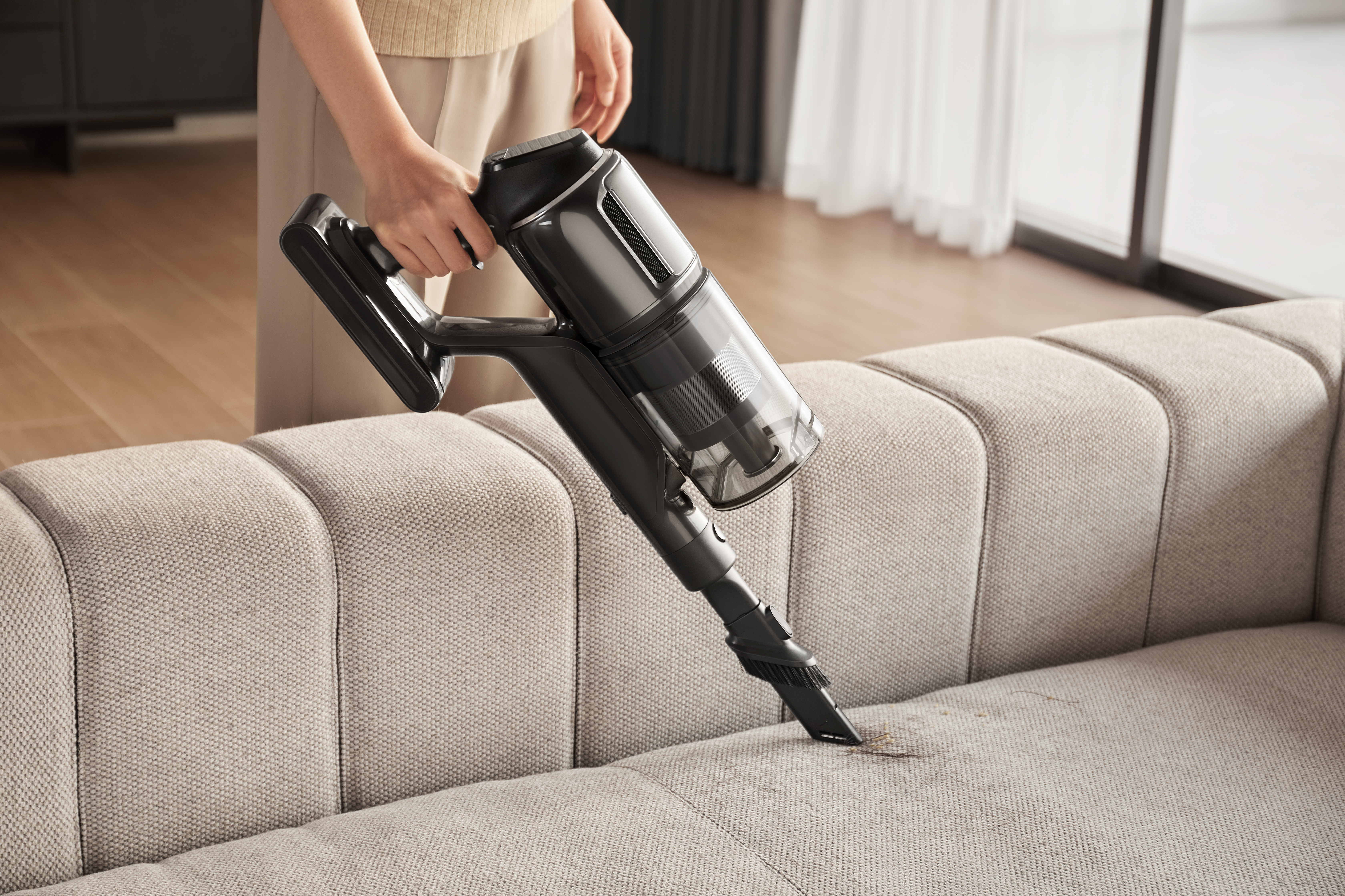 Osoba odkurza beżową sofę czarnym odkurzaczem. Brud jest widoczny na sofie.