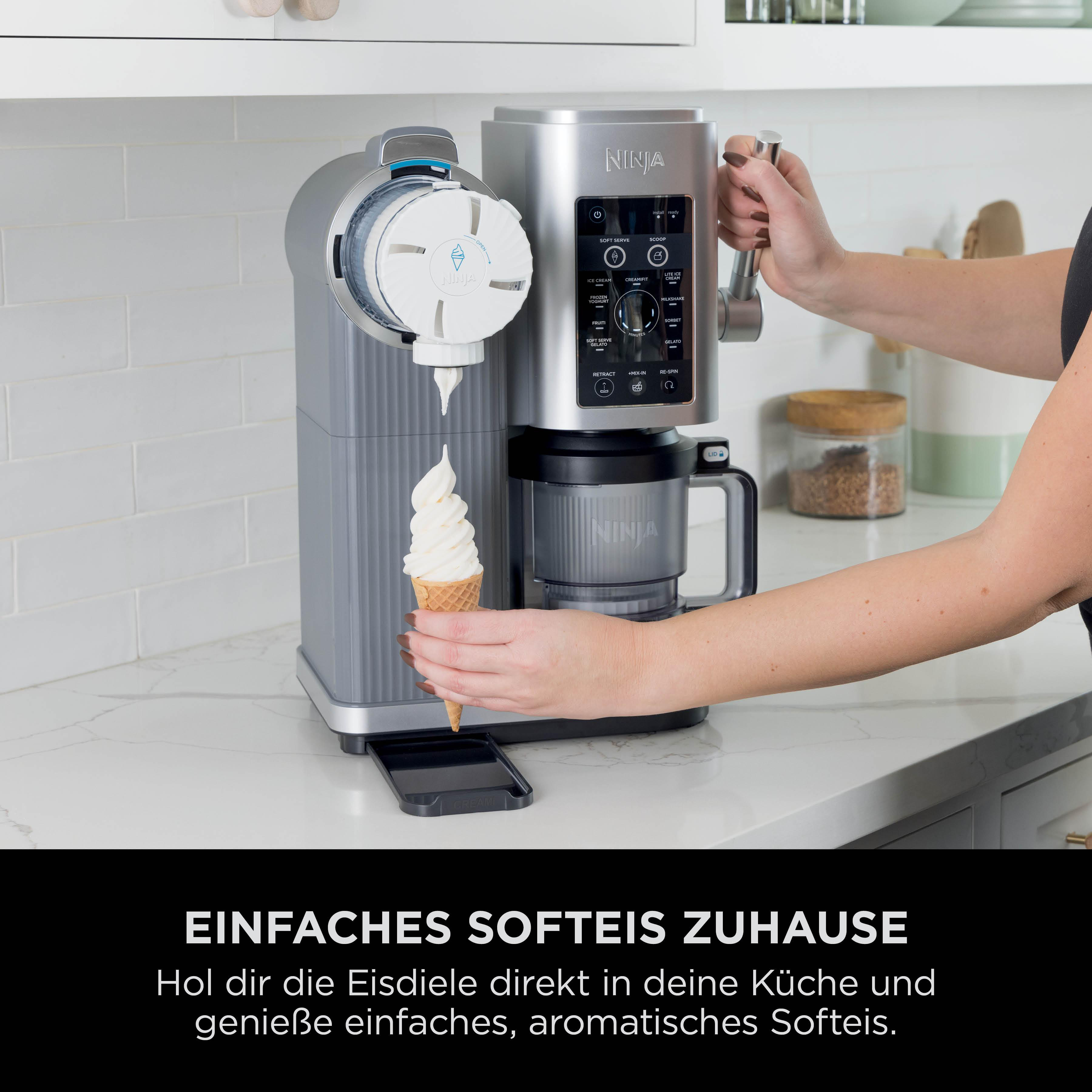 Ein silberner Ninja-Eismaschine gibt Softeis in einen Kegel ab. Die Hand einer Person hält den Kegel. Der Hintergrund ist eine weiße Küche.