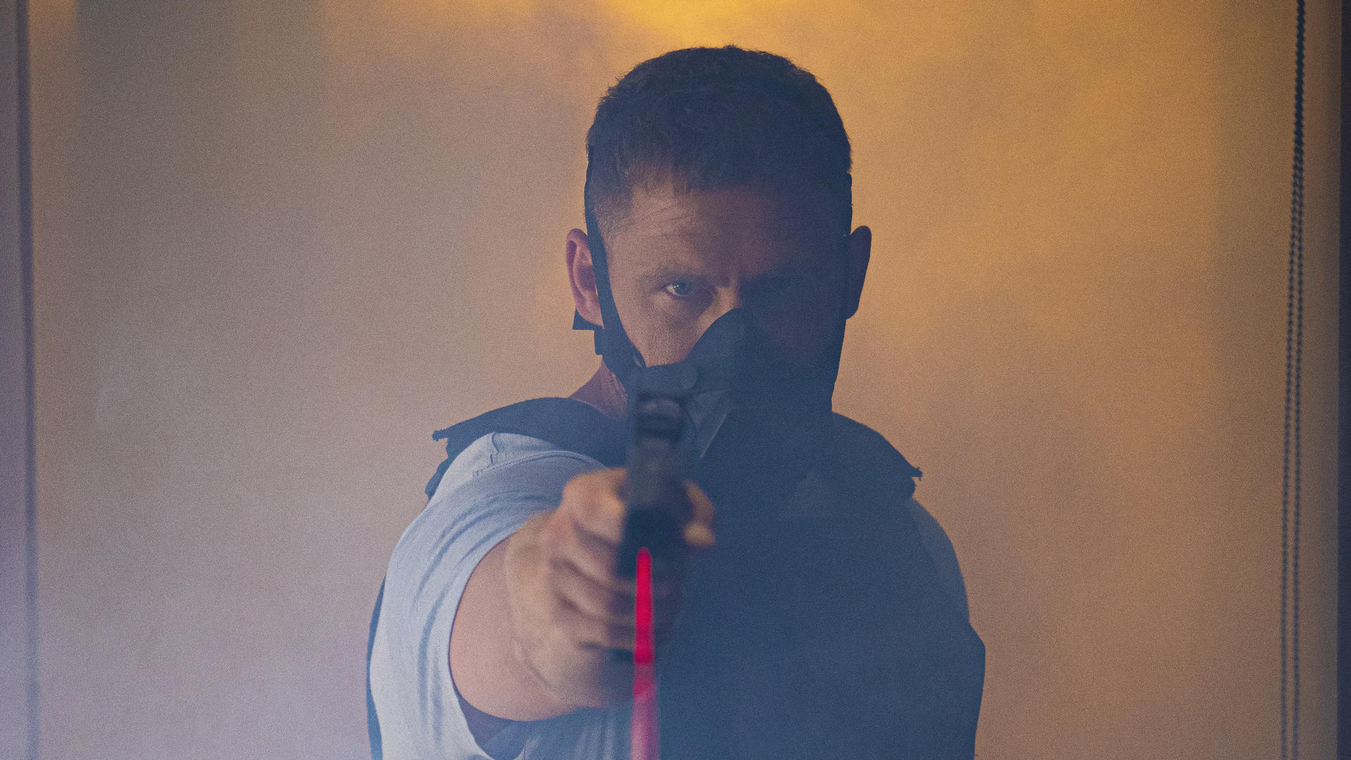 Ein Mann mit Maske zielt mit einer Waffe. Er befindet sich in einem rauchigen Raum und trägt ein hellblaues Hemd und eine dunkle Weste.