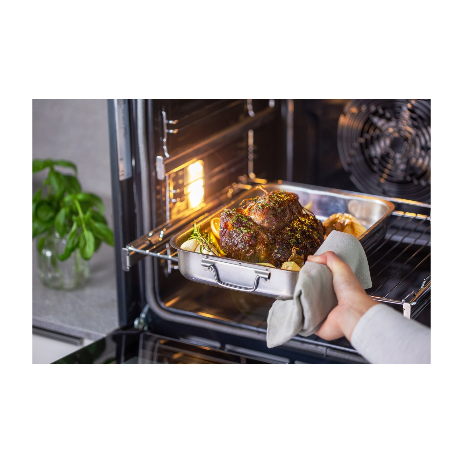 Una persona toglie un arrosto dal forno. L'arrosto è in una teglia di metallo con limoni e aglio. Erbe verdi sono vicine in un vaso di vetro.