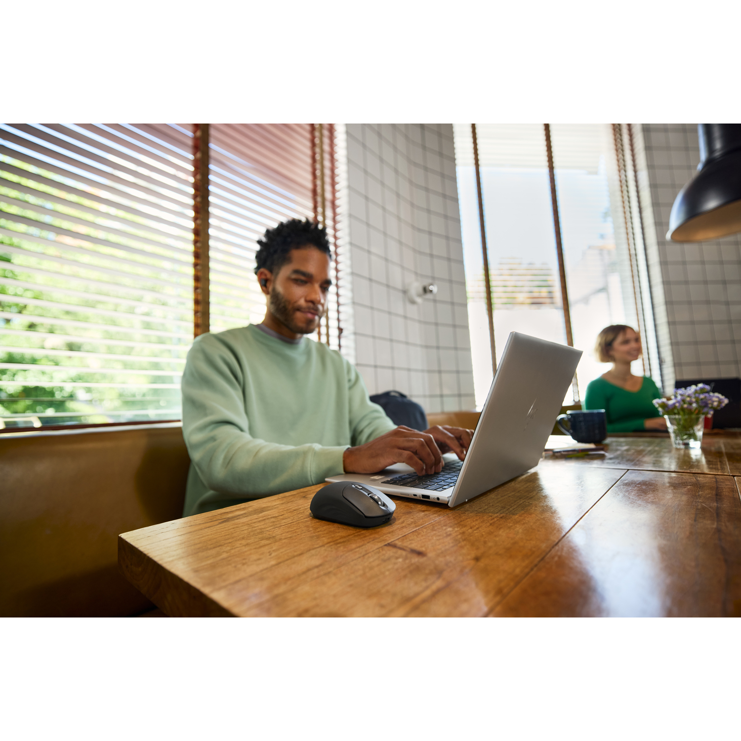Un homme tape sur un ordinateur portable à une table. Une souris d'ordinateur est à côté de l'ordinateur portable. Une autre personne est en arrière-plan.