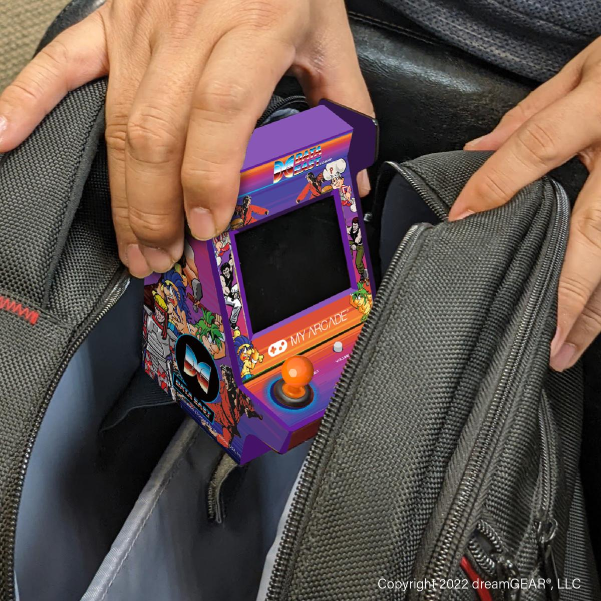 Des mains tiennent une mini-borne d'arcade, la plaçant dans un sac noir. Le jeu est violet, avec un joystick et un écran.