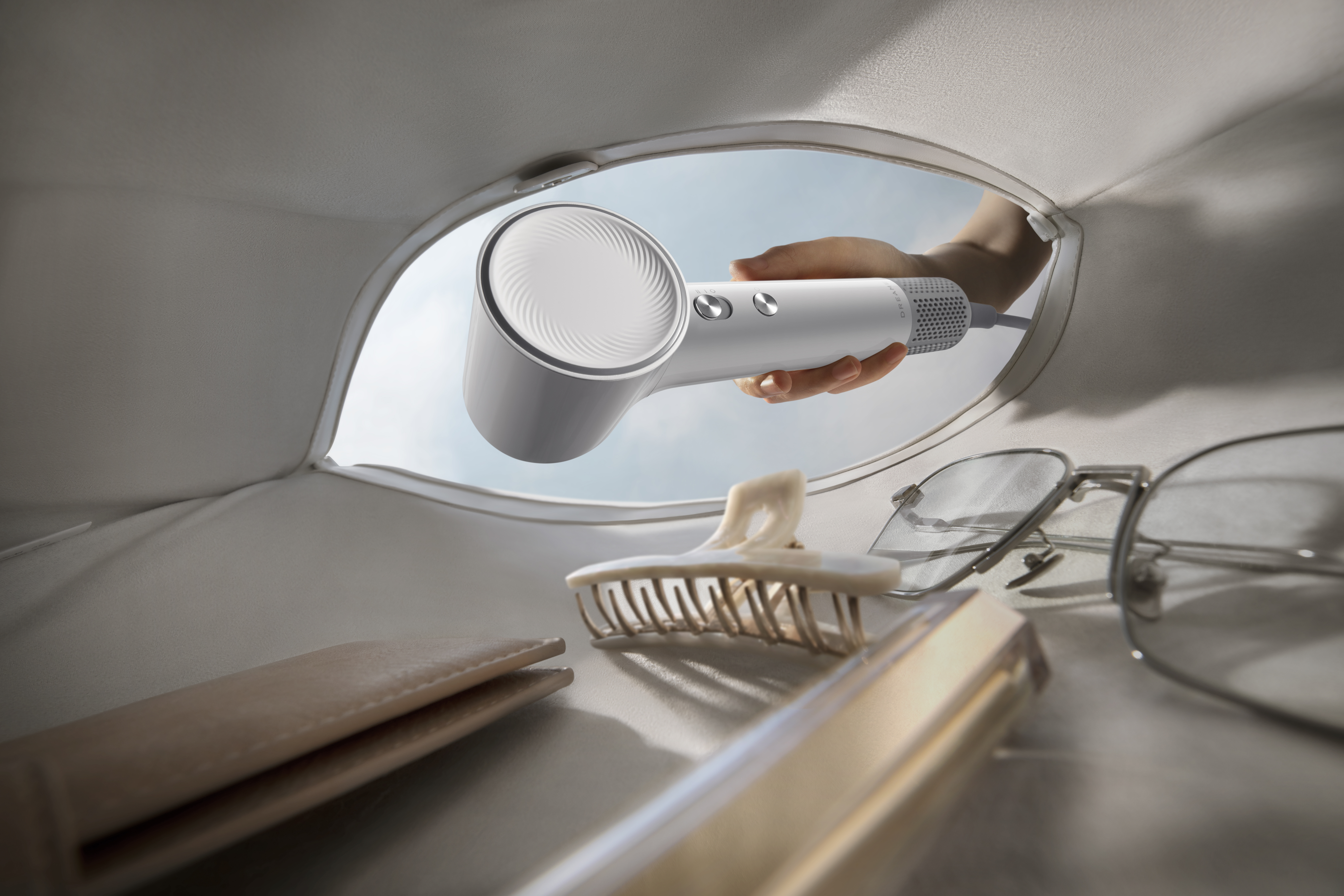 Ręka trzyma biało-szarą suszarkę do włosów, wyłaniającą się z jasnej torby. W torbie znajdują się akcesoria, takie jak okulary i spinka do włosów.