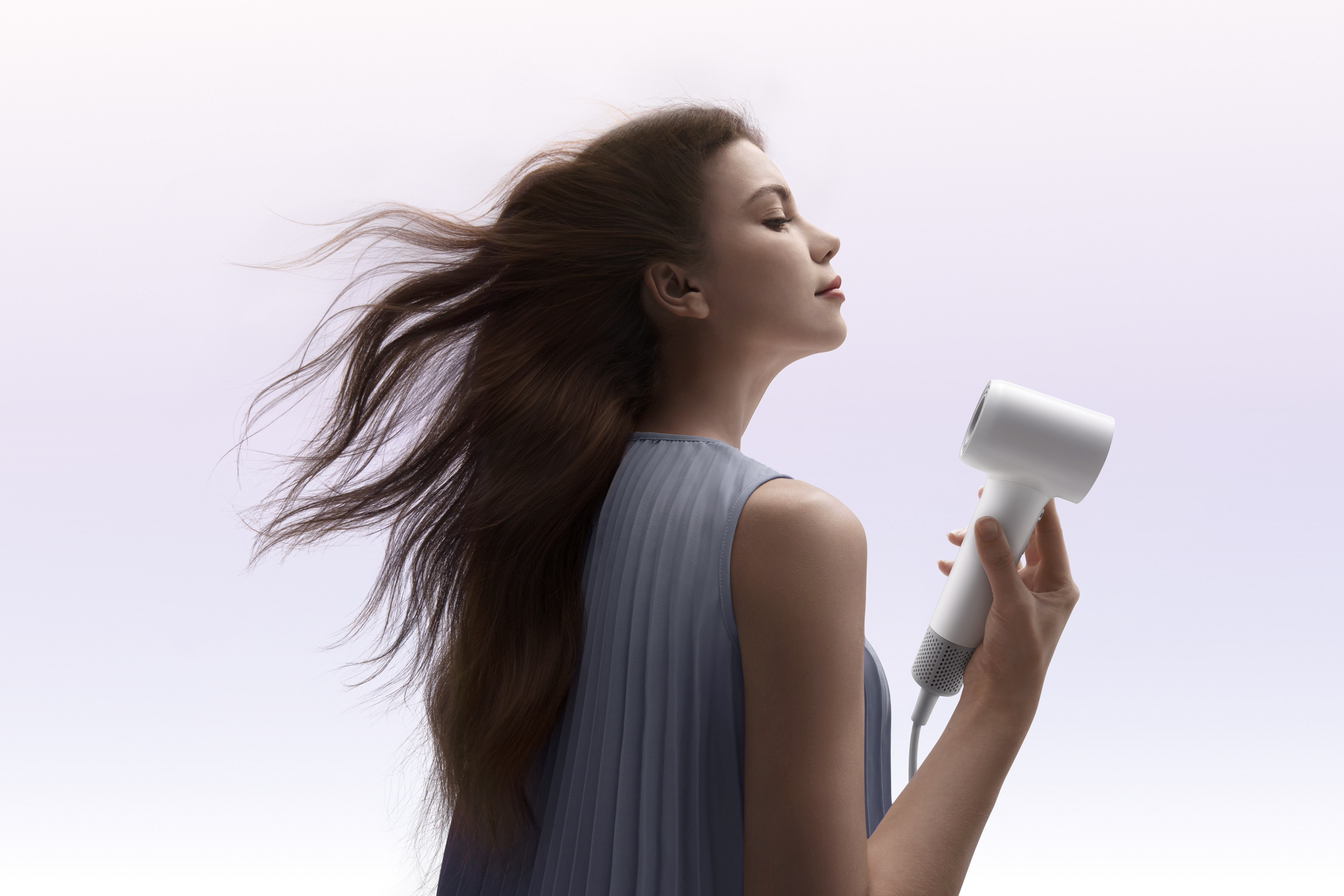 Kobieta z rozwianymi brązowymi włosami trzyma białą suszarkę do włosów. Ma na sobie niebieską sukienkę, oczy zamknięte, na białym tle.