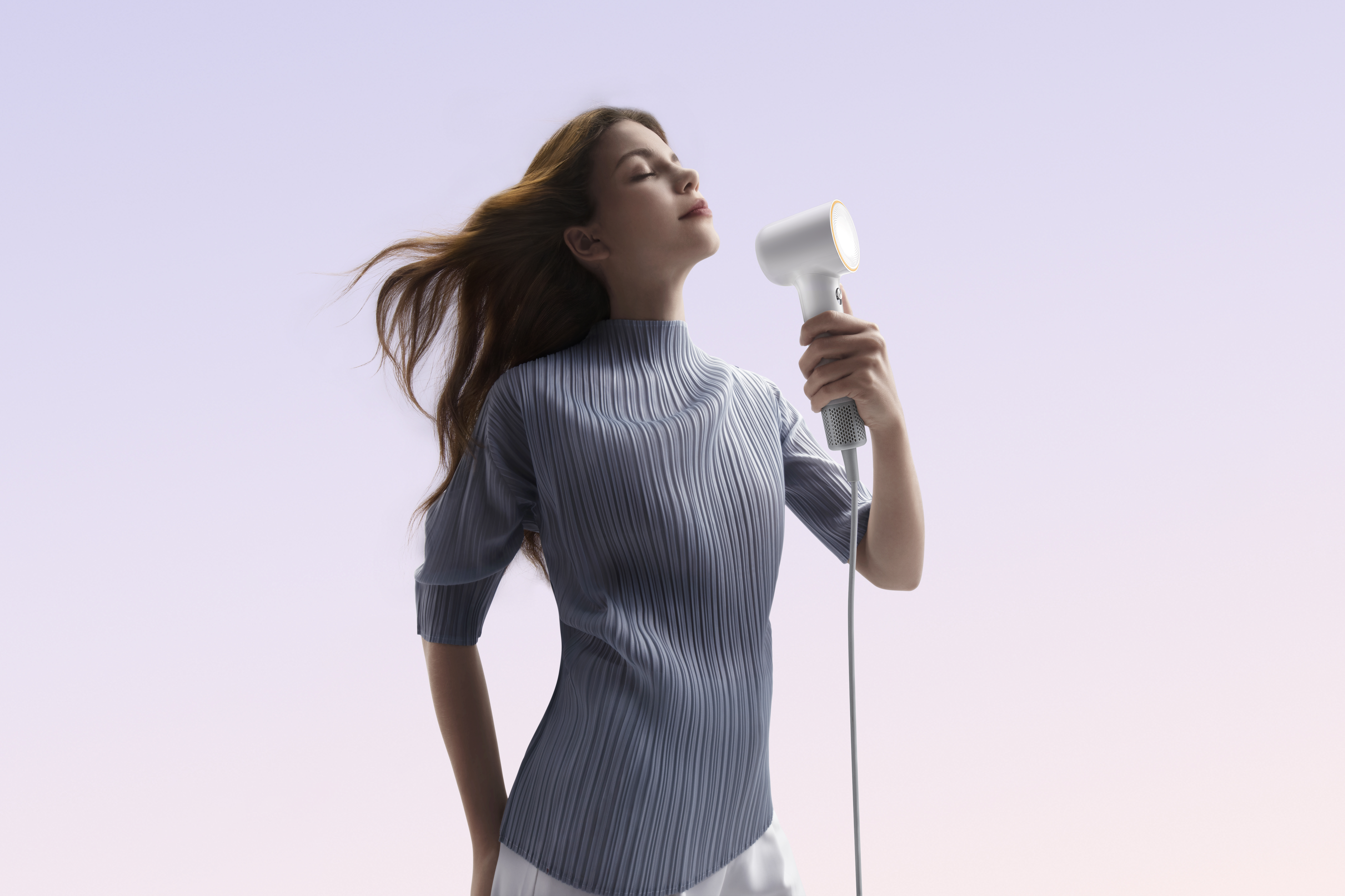 Kobieta z rozwianymi włosami trzyma suszarkę, oczy zamknięte. Ma na sobie niebieski top na gradientowym tle.