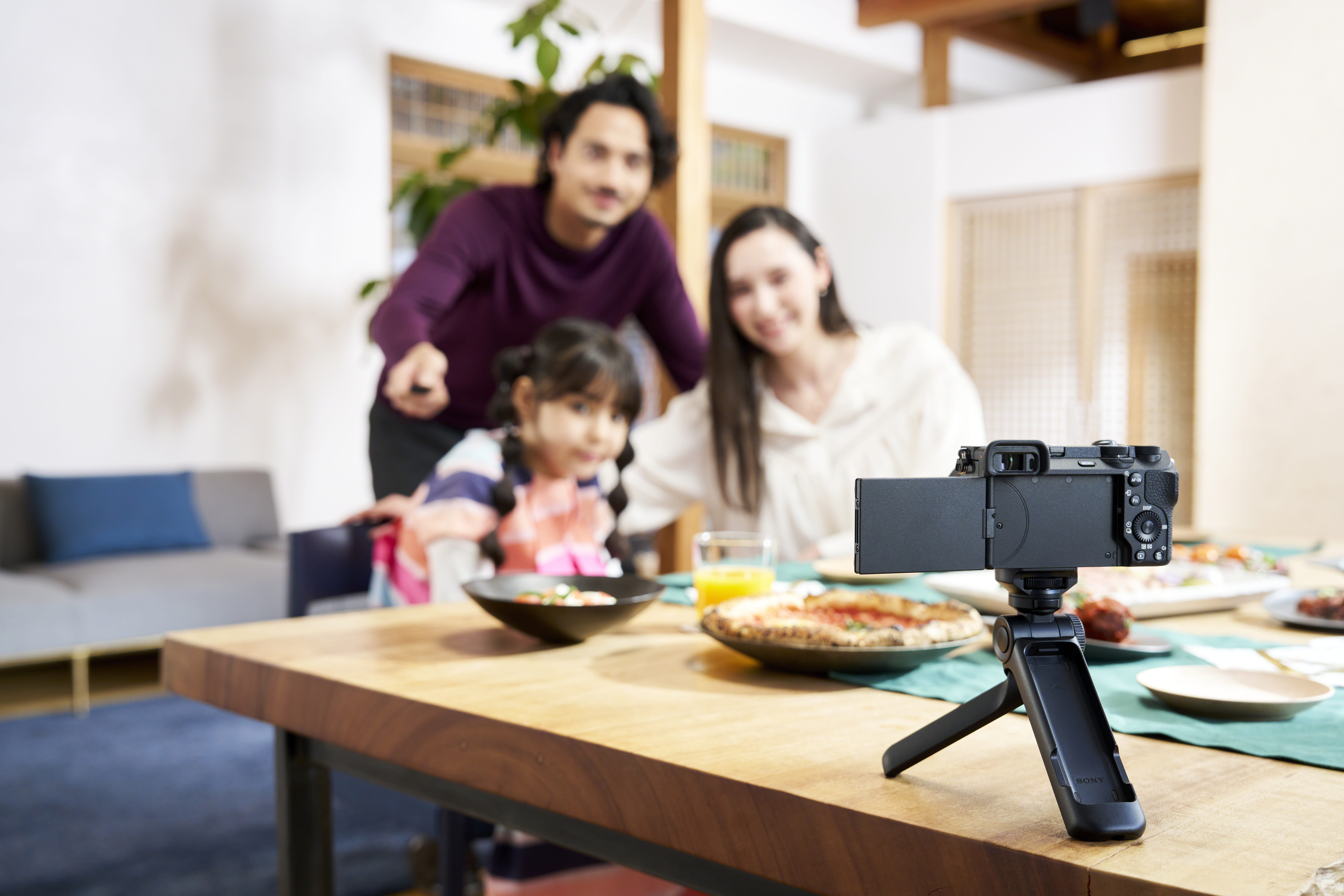 Familia en una mesa con comida, una cámara está enfocada. Un hombre señala. Una niña y una mujer sonríen.