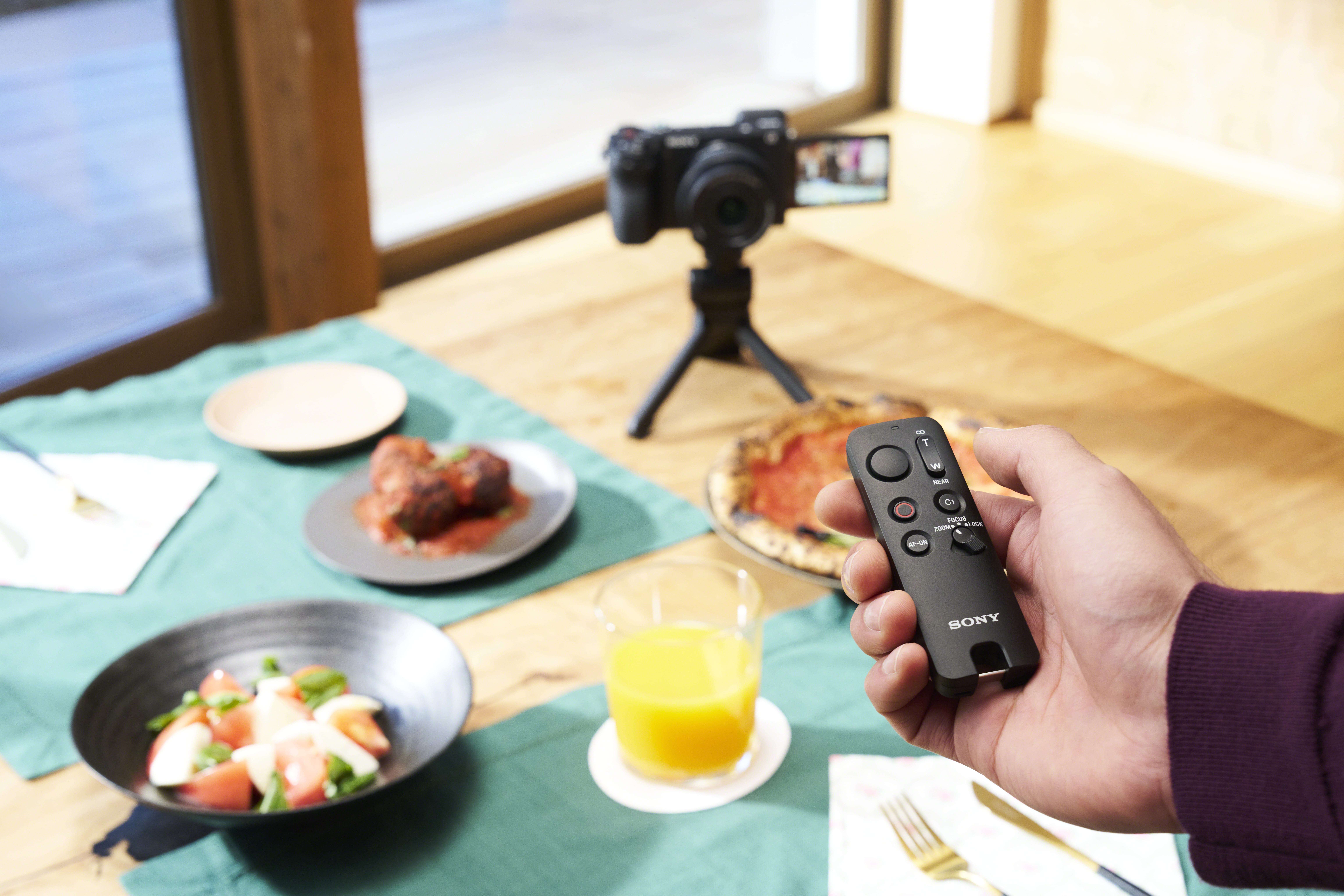 Una mano sostiene un mando a distancia Sony. En el fondo: cámara, comida y un vaso de zumo en una mesa.