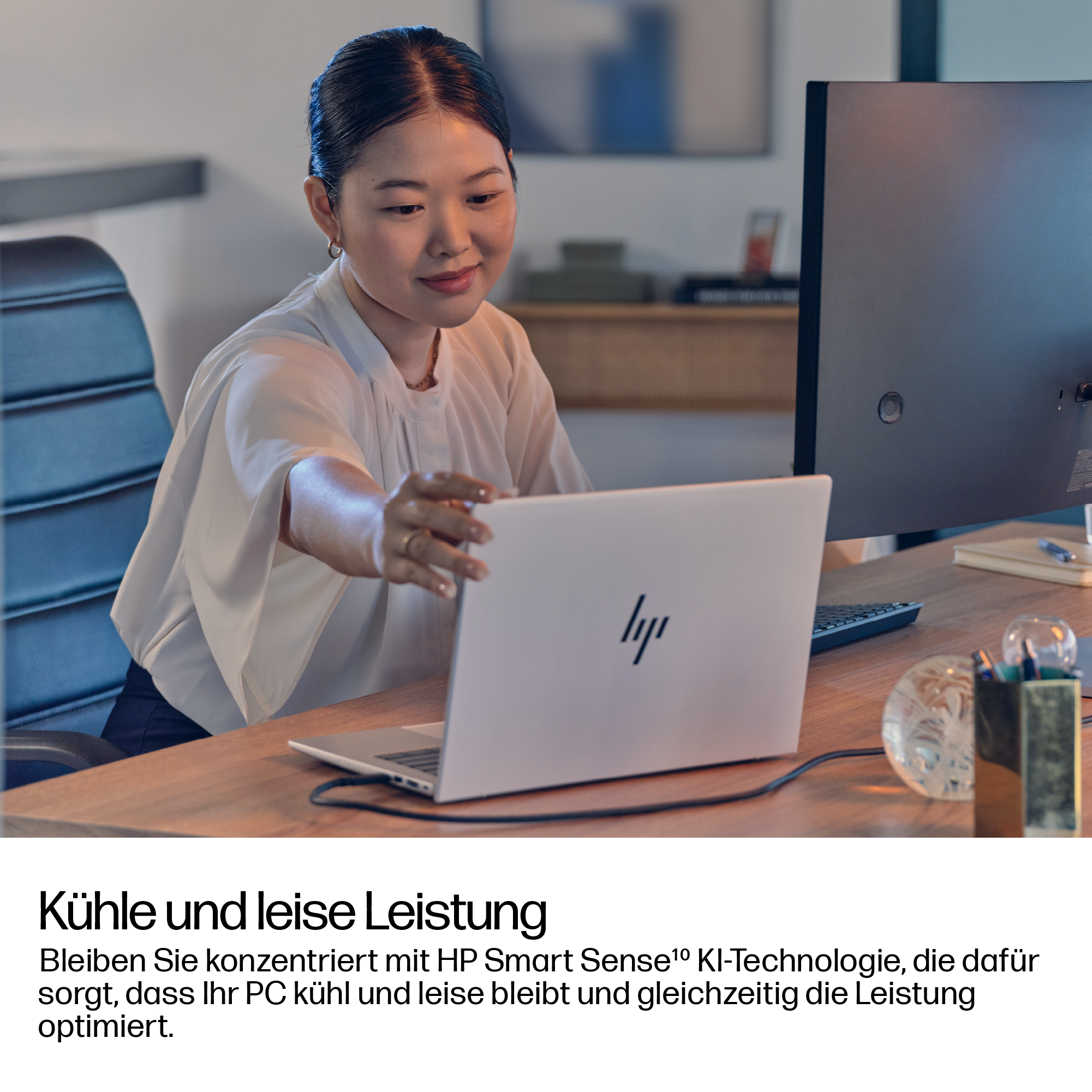 Eine Frau benutzt einen weißen HP-Laptop an einem Schreibtisch. Ein Monitor und Büromaterial sind im Hintergrund zu sehen.