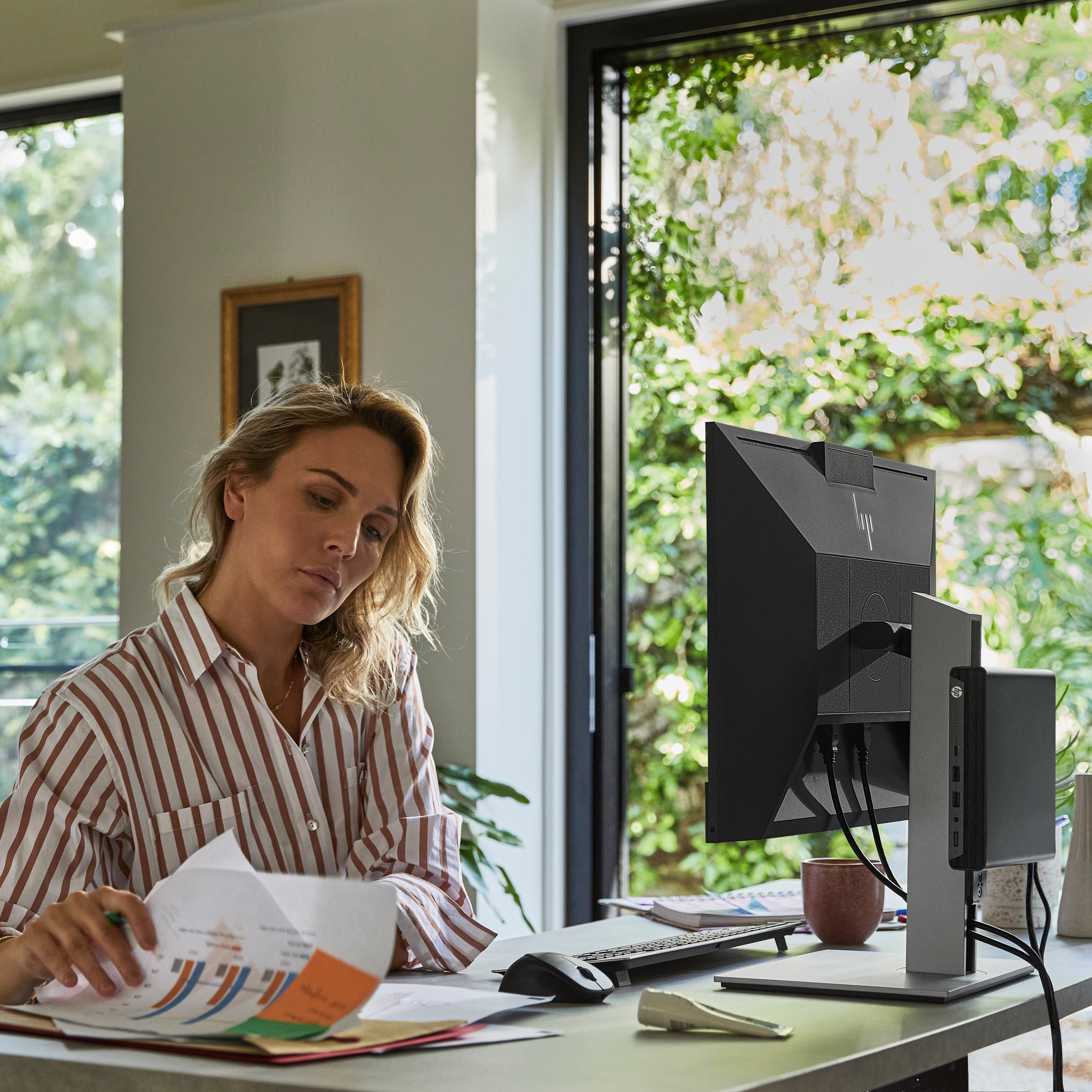 Frau am Schreibtisch prüft Papiere. Computermonitor und Zubehör sind vorhanden. Grüne Pflanzen sind vor einem Fenster sichtbar.
