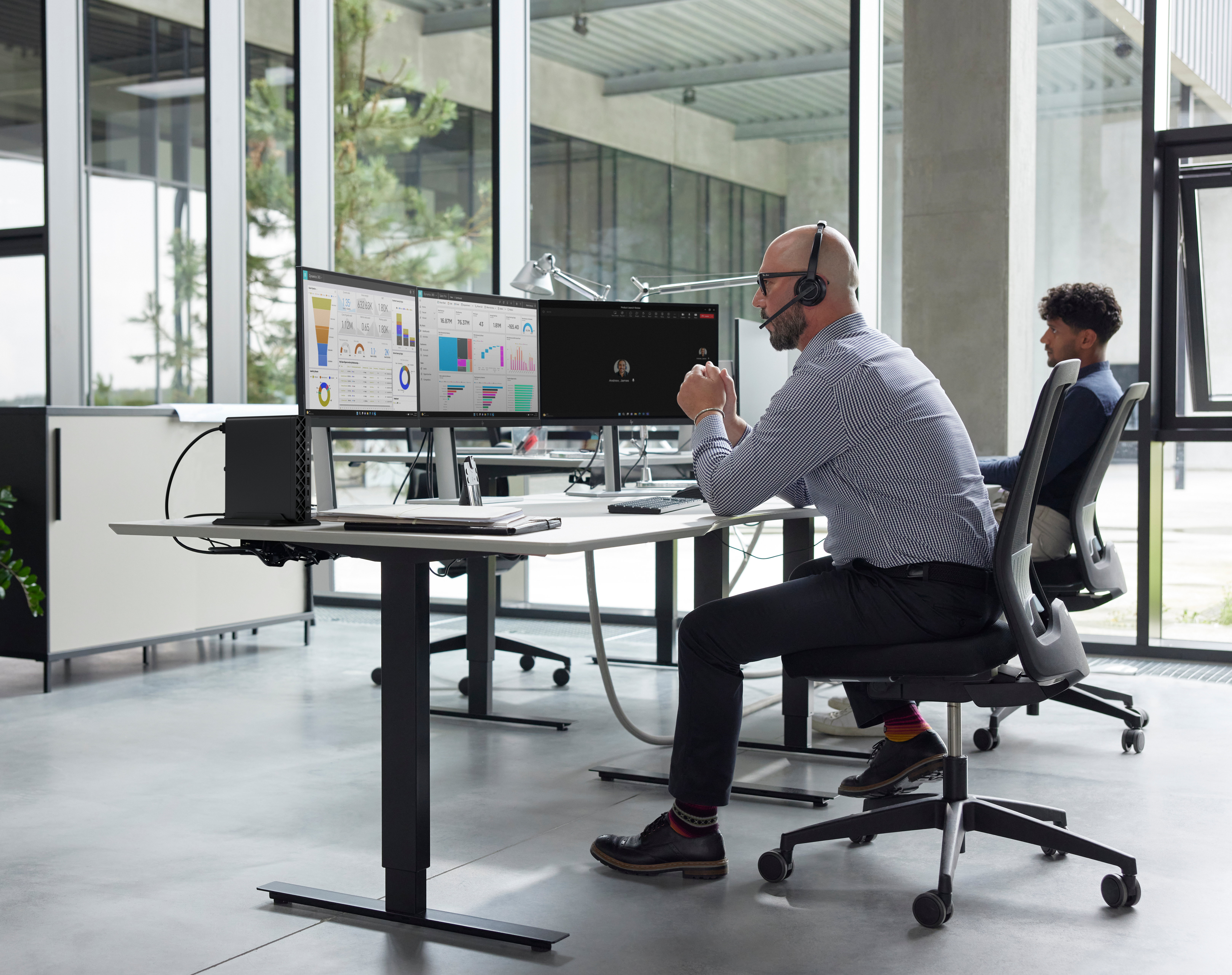 Zwei Männer an Schreibtischen. Einer trägt ein Headset und schaut auf Monitore. Ein anderer sitzt im Hintergrund.
