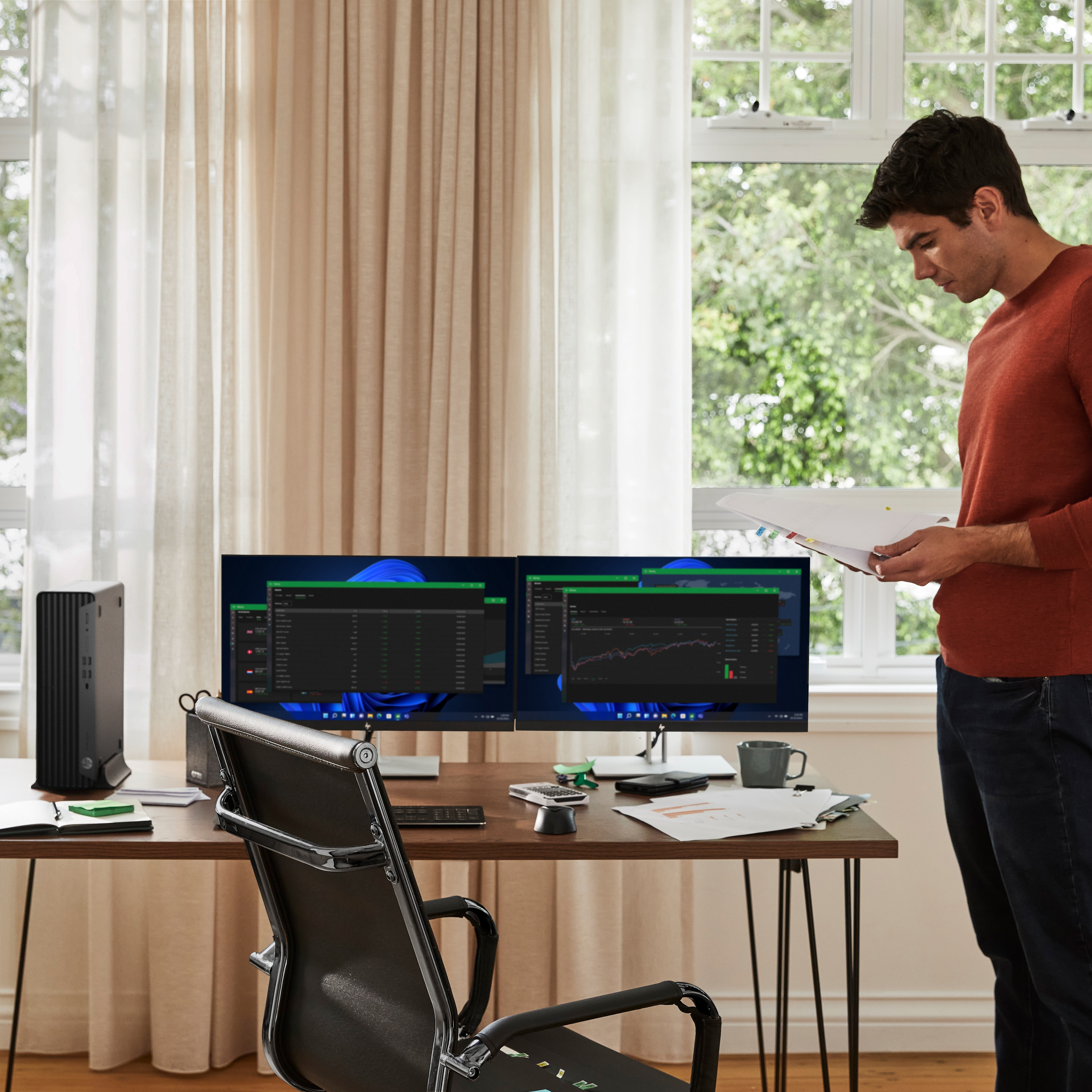 Mann in orangefarbenem Pullover betrachtet Papiere im Homeoffice. Monitore zeigen Grafiken. Auf dem Schreibtisch stehen ein Computer, eine Tasse und ein Stuhl.