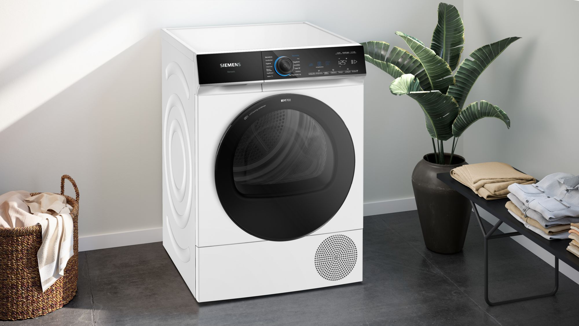 Ein weißer Siemens-Trockner mit schwarzer Tür. Rechts eine Pflanze im Topf. Kleidung liegt auf einer schwarzen Bank.