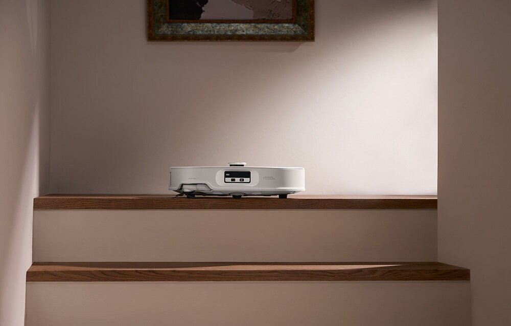 Biały odkurzacz automatyczny stoi na drewnianym stopniu. Nad nim obraz w ramie wisi na jasnej ścianie.