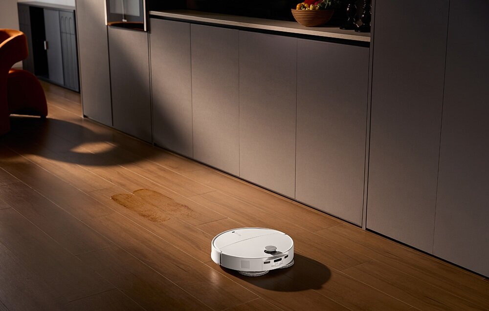 Biały robot odkurzający na drewnianej podłodze. Widoczna plama wody. Szafki kuchenne w tle.