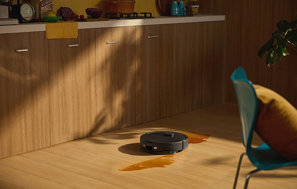 Robot sprząta podłogę w kuchni z rozlanym płynem. W tle krzesło i drewniane szafki.