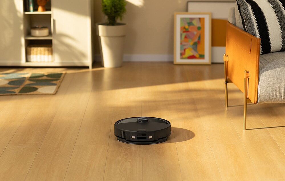 Czarny robot odkurzający na drewnianej podłodze. W tle sofa, obraz i roślina.