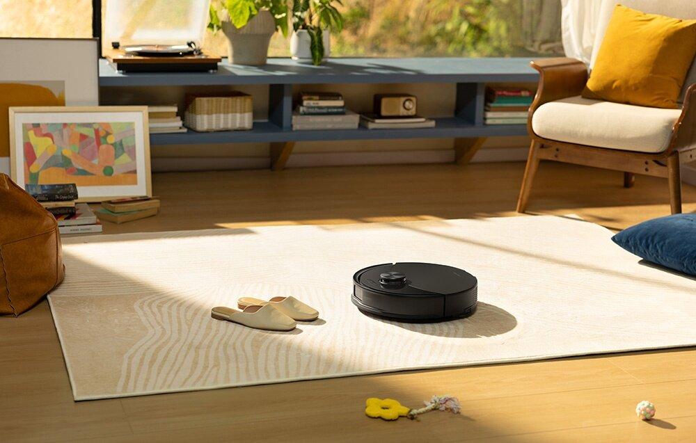 Scena w salonie. Czarny odkurzacz robot na dywanie. Widoczne są również krzesło, kapcie i obraz.