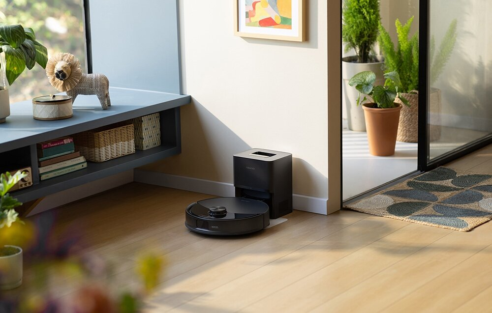 Czarny robot odkurzający i stacja ładująca na drewnianej podłodze. W tle statua lwa, rośliny i obraz.