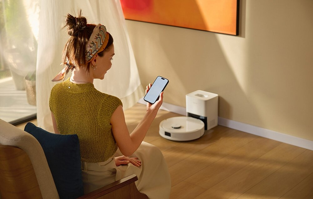 Kobieta trzyma telefon, patrząc na robota odkurzającego. Stacja ładująca i obraz są w tle.
