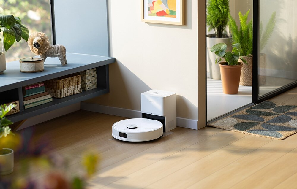 Biały odkurzacz automatyczny na drewnianej podłodze. Obok biała stacja ładująca. Rośliny i posąg lwa na półce.