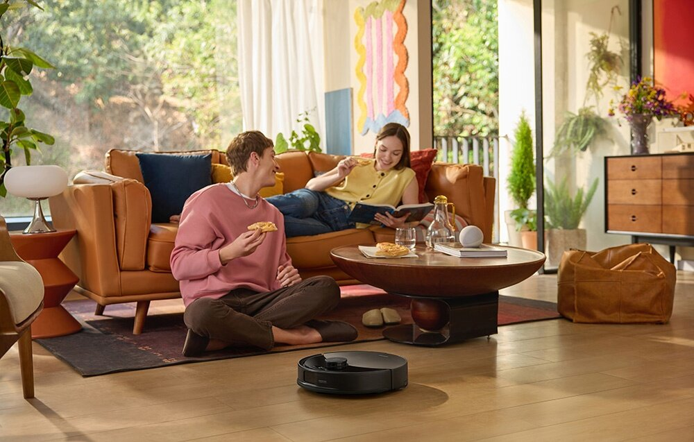Para relaksuje się w salonie, jedząc pizzę. Robot sprzątający stoi na podłodze. Brązowa skórzana sofa w tle.