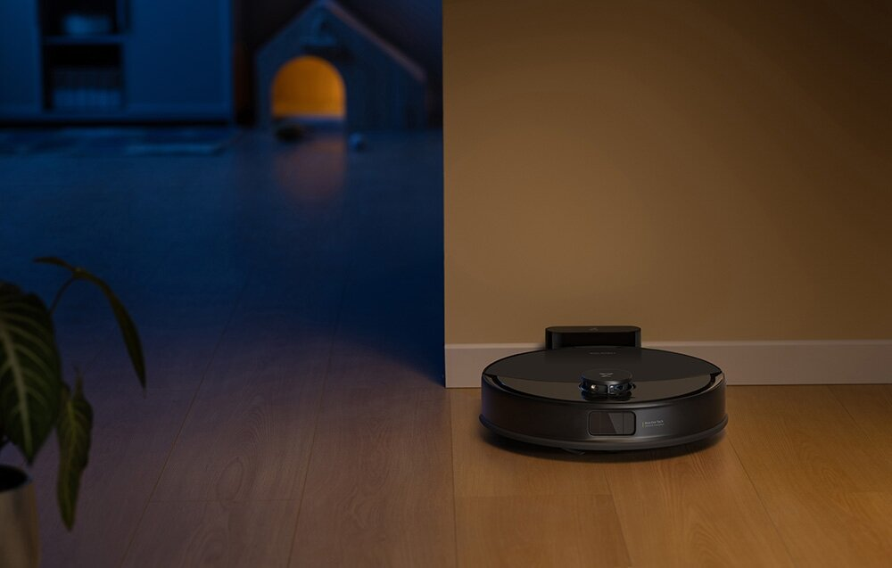 Czarny odkurzacz robot na drewnianej podłodze stoi obok beżowej ściany. Lewa strona jest ciemna, z domkiem dla zabawek.