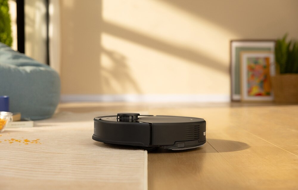 Czarny odkurzacz robot na jasnej drewnianej podłodze. Niebieski worek do siedzenia, ramki i roślina w tle.