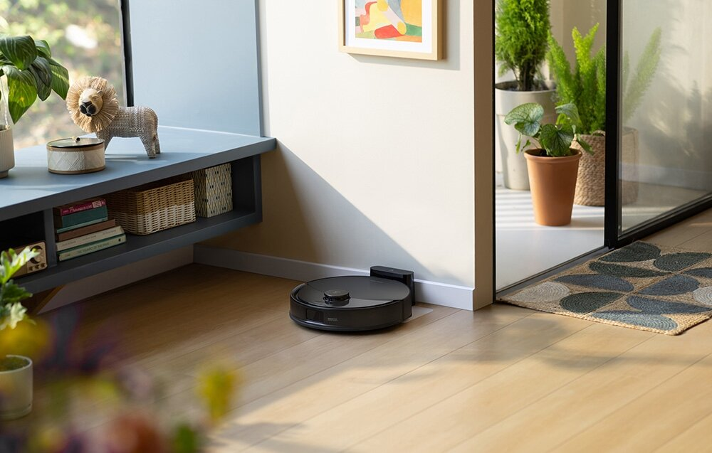 Czarny odkurzacz automatyczny stoi na drewnianej podłodze. Posąg lwa stoi na półce. Widoczne są również rośliny i dywan.