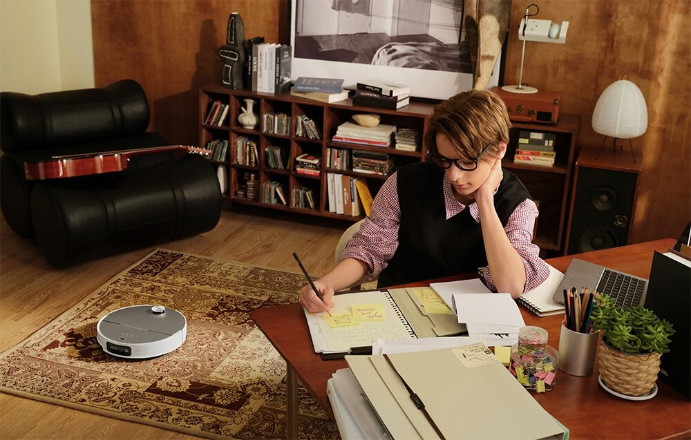 Osoba w okularach siedzi przy biurku i pisze. Na scenie dywan z robotem sprzątającym, gitara na czarnym krześle i regał z książkami.