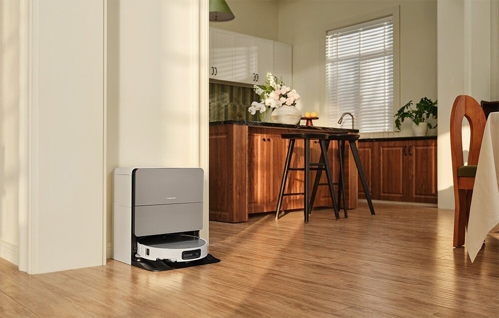 Robot odkurzający stoi w kuchni. Drewniane szafki i podłogi. Po prawej stronie obrazu znajduje się stół.
