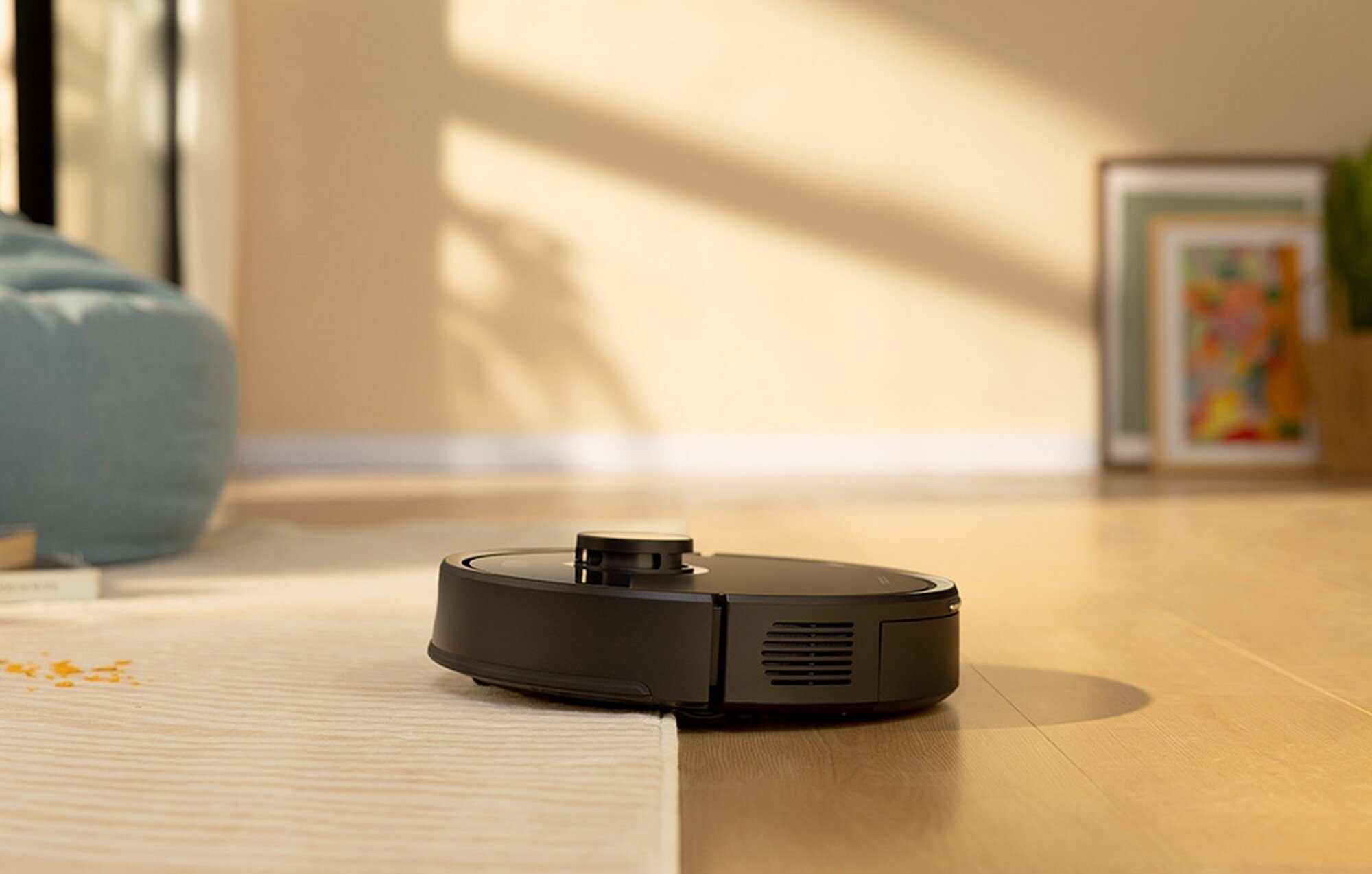 Czarny odkurzacz robot na dywanie, z drewnianą podłogą w tle. Widoczne są również ramka na zdjęcia i roślina.
