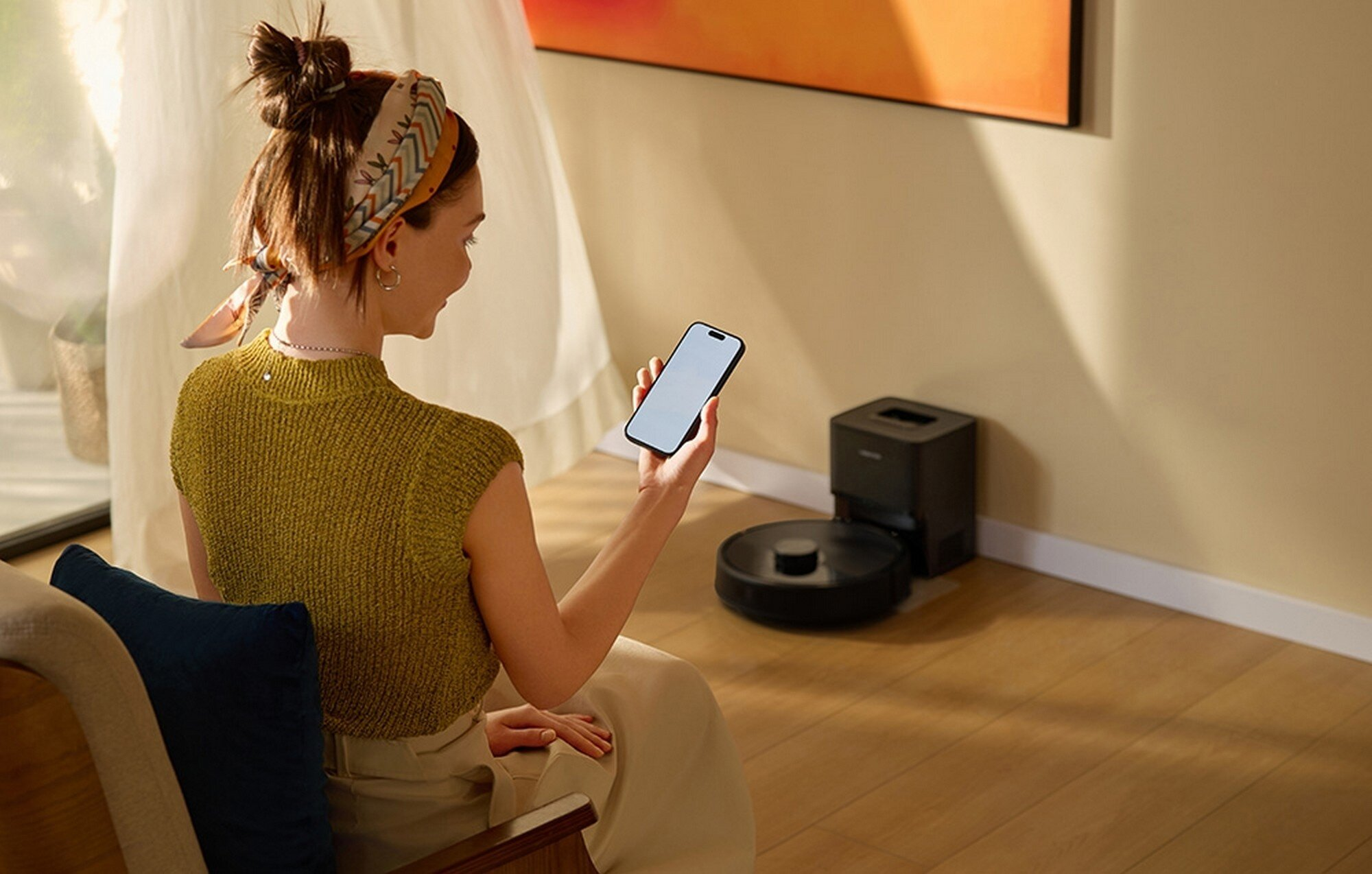 Kobieta trzyma telefon, patrząc na robota odkurzającego. Stacja ładująca jest w pobliżu. Ma na sobie żółtą koszulę i szalik.