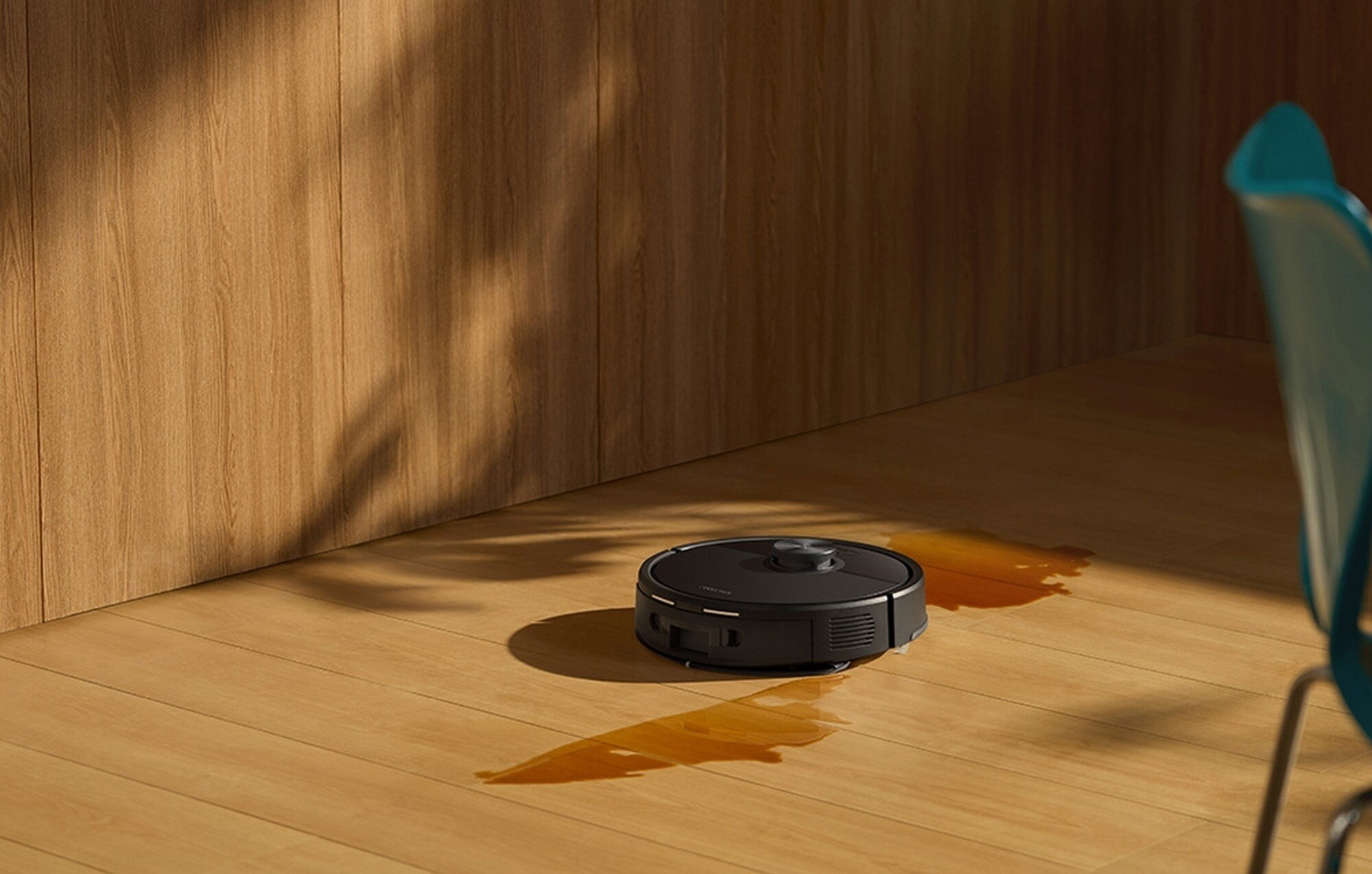 Czarny odkurzacz robot na drewnianej podłodze. Obok niego plama. Drewniana ściana i turkusowe krzesło w tle.