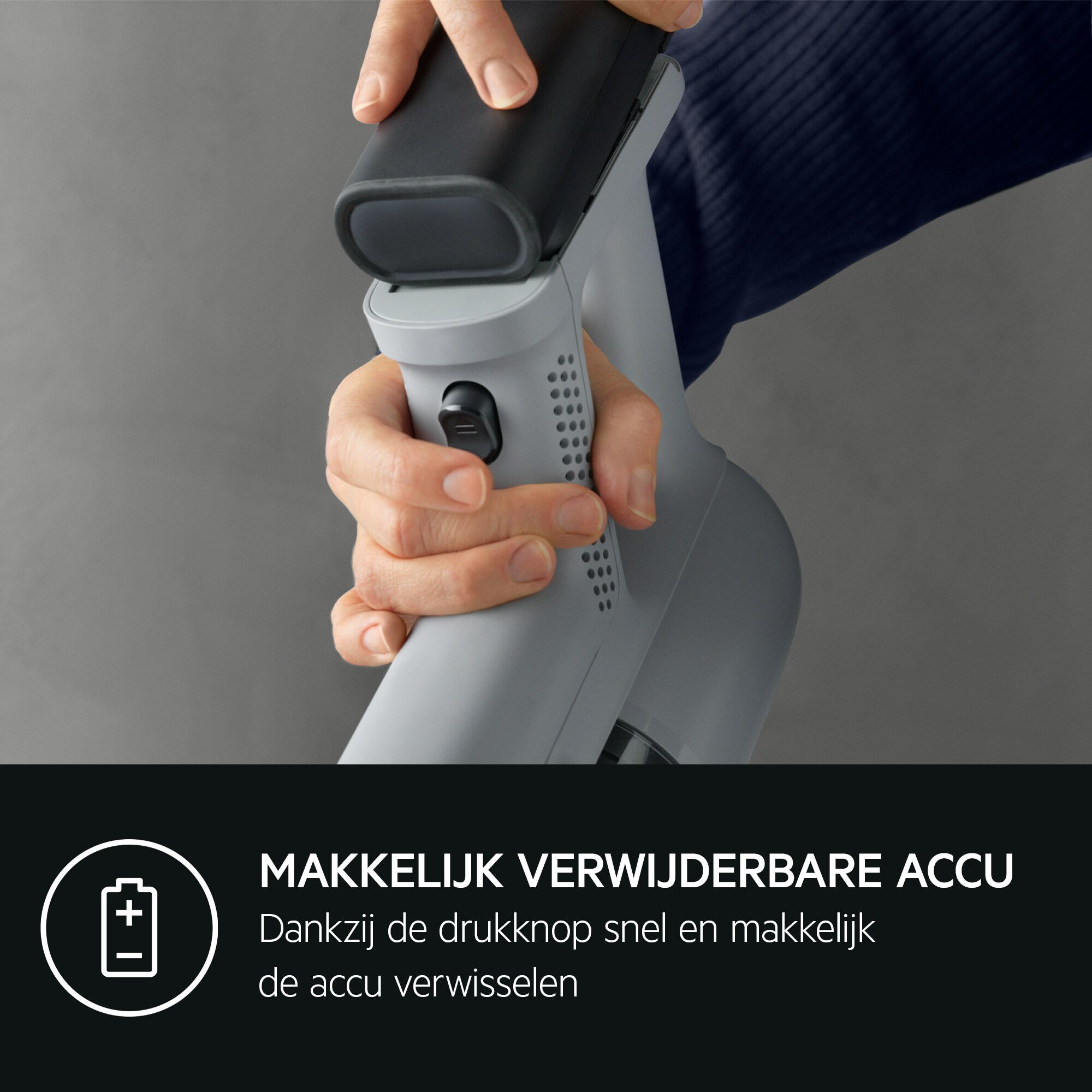 Hand holding a black battery pack, removing it from a gray vacuum cleaner. The background is gray.