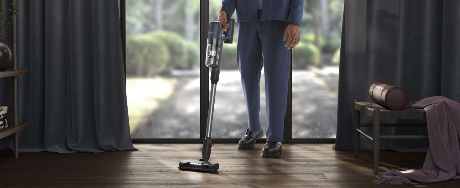 Person in blue suit holding a vacuum cleaner, standing on a wooden floor. Background has curtains and a window.